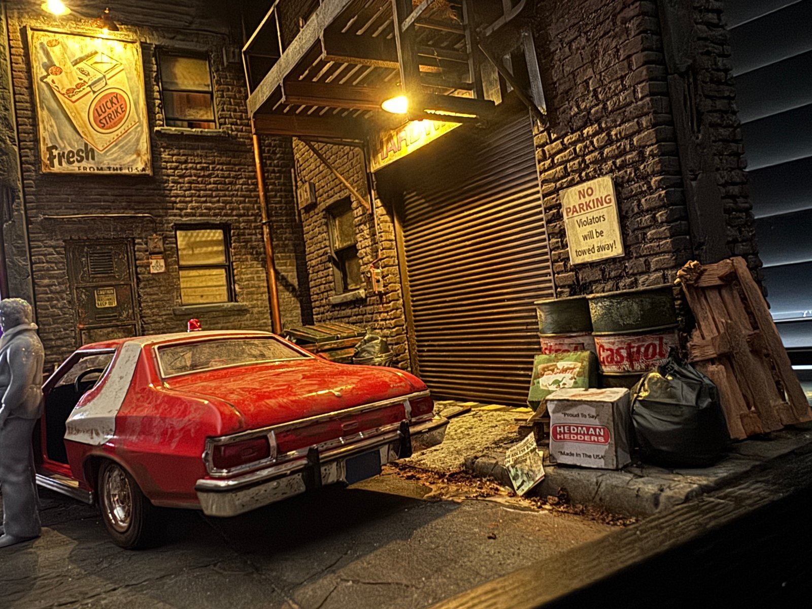 Rear three-quarter view of the weathered 1:24 scale red Ford Gran Torino in the Starsky and Hutch diorama, surrounded by detailed street debris including Castrol oil drums, wooden crates, and a Hedman Hedders box on a realistic cobblestone base.