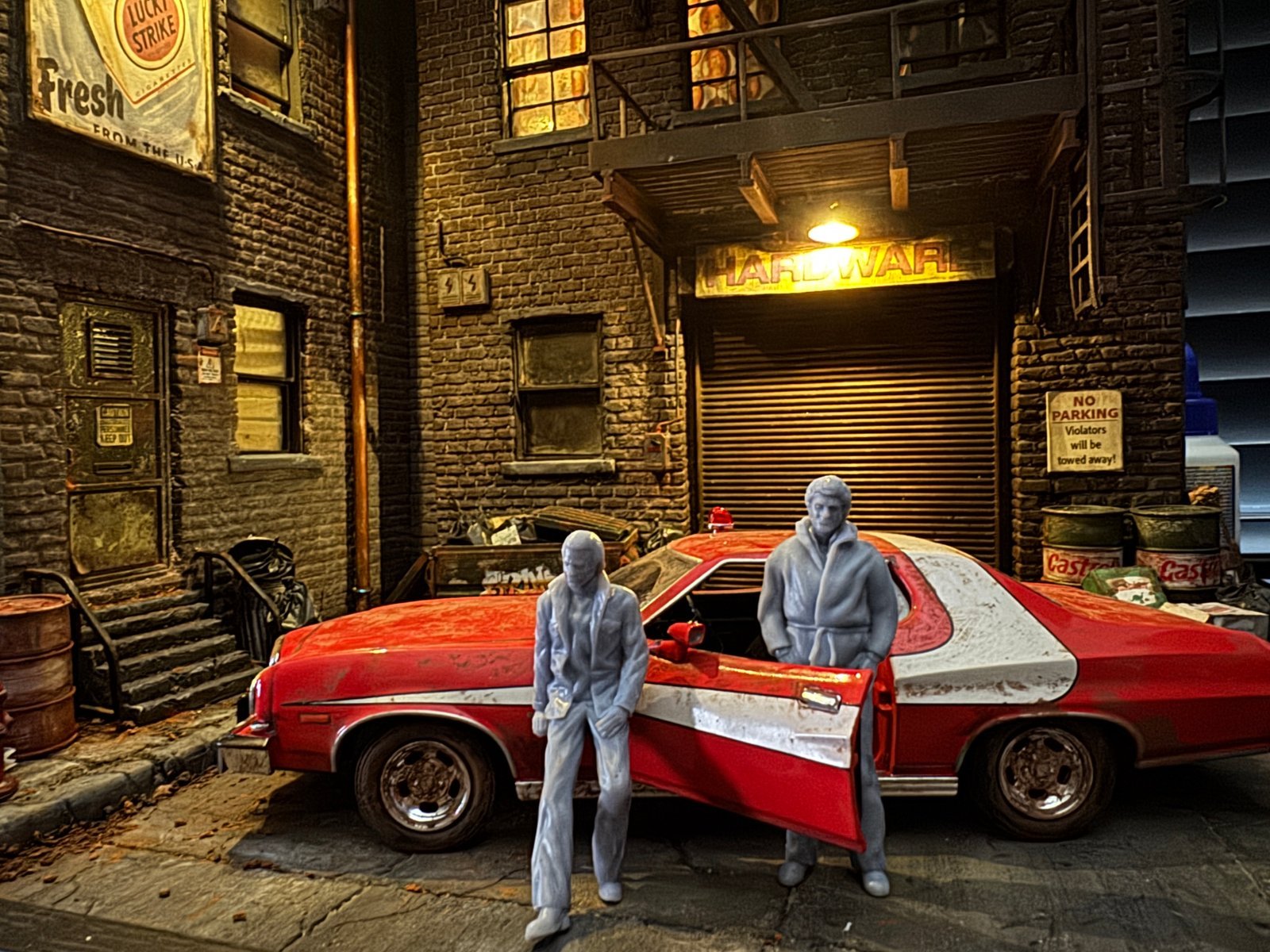 Starsky and Hutch 1:24 scale diorama featuring two unpainted resin figures standing in front of the iconic red and white Ford Gran Torino, set against a gritty 1970s urban brick alley backdrop with a Hardware store shutter door.