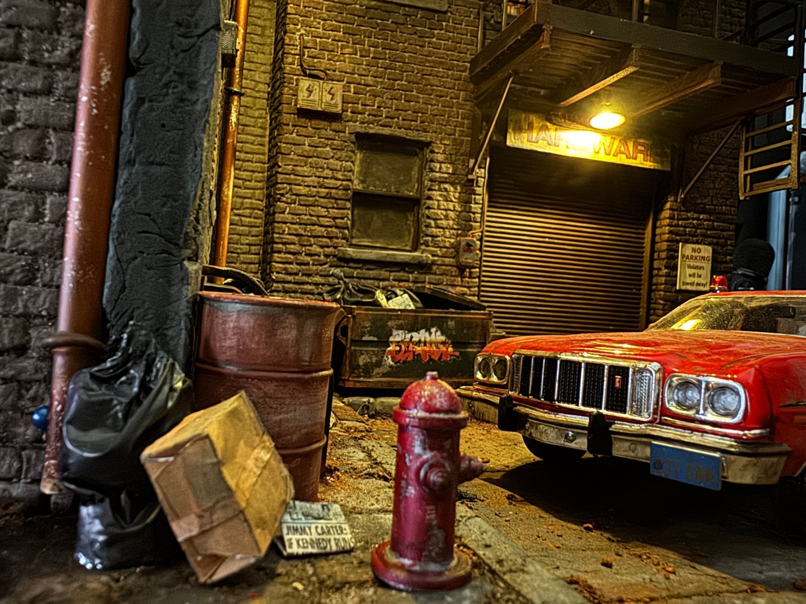 Low-angle close-up of the Starsky and Hutch 1:24 scale diorama street scene featuring a weathered red fire hydrant, rusted steel drum, cardboard box, and Jimmy Carter newspaper prop in the foreground, with the red Gran Torino and Hardware store in the background.